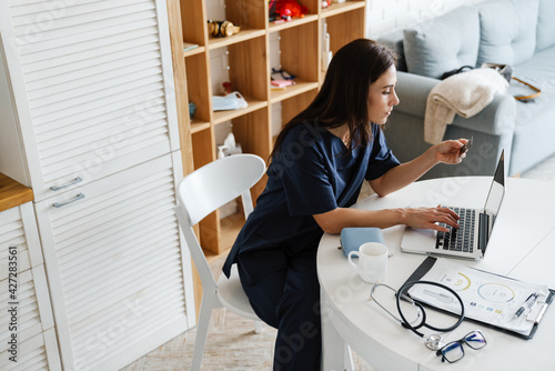 Focused woman doctor using credit card and laptop while sitting at home