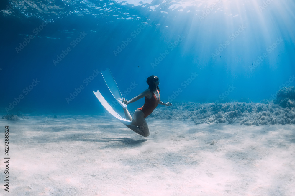 Woman freediver in swimsuit with fins glides underwater over sand in ocean.