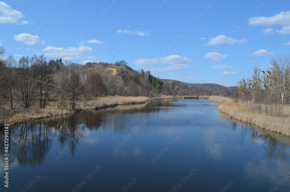 Beautiful landscape with a wide blue river and high hill on the horizon. Reflection of the sky and clouds in the water. Early spring nature in sunny warm weather.