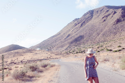 young woman with long brunette hair wears a hat and a floral dress walking along a path in a dry and hilly yellow spot in the late afternoon