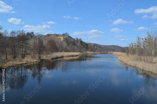 Beautiful landscape with a wide blue river and high hill on the horizon. Reflection of the sky and clouds in the water. Early spring nature in sunny warm weather.