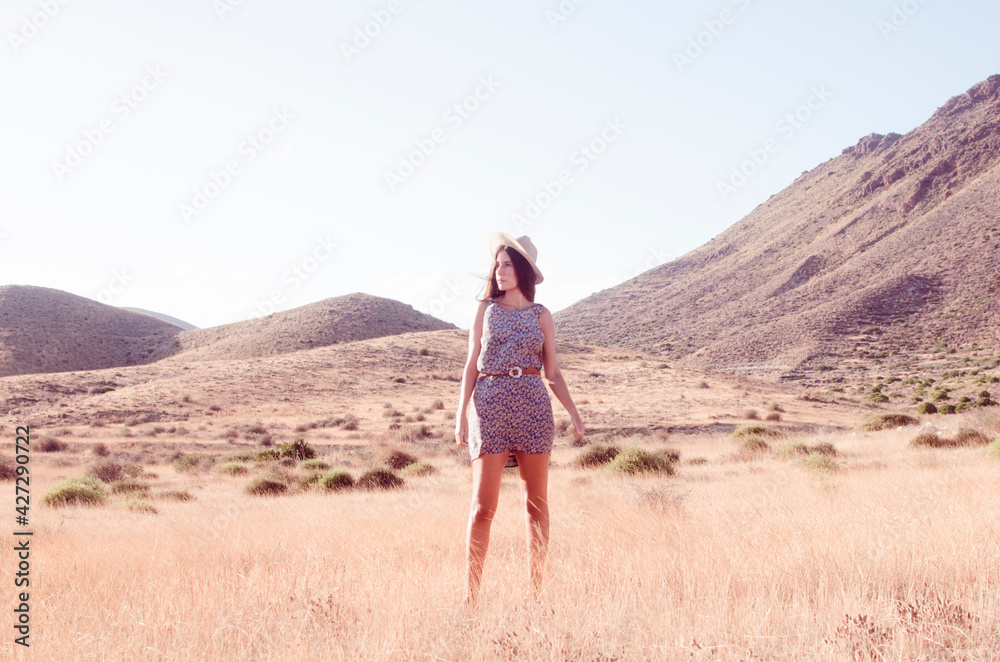 Young woman with long hair posing wears hat and  floral dress in a dry hilly meadow in the late afternoon