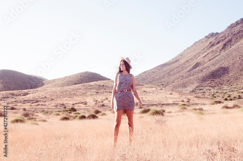 Young woman with long hair posing wears hat and floral dress in a dry hilly meadow in the late afternoon