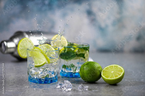 Mojito cocktail with splash and drops. Summer alcoholic cocktail mojito with rum, mint, lime and ice, bar tools on concrete gray blue background