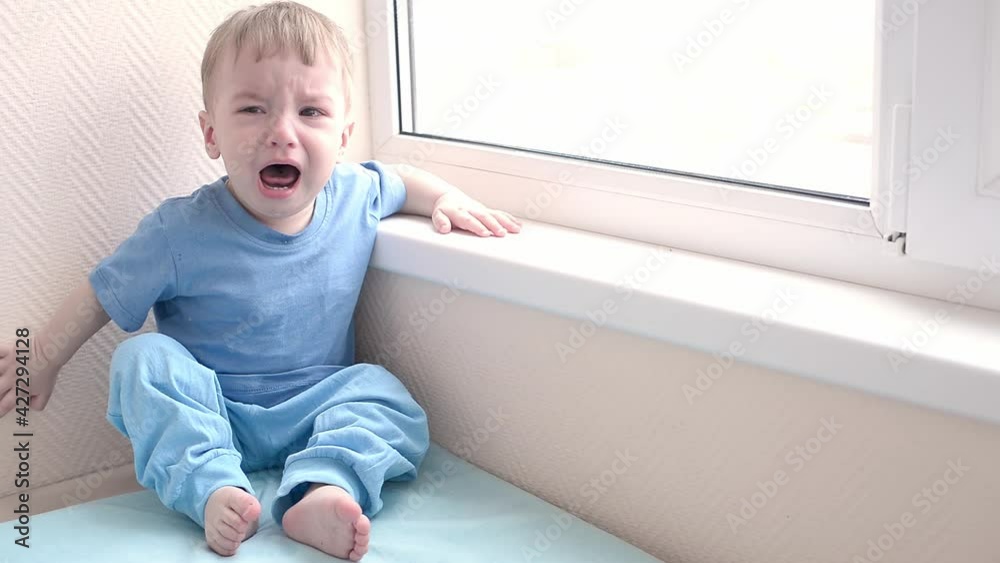 little toddler boy sitting by the window and crying loudly. little toddler boy sitting by the window and crying loudly.