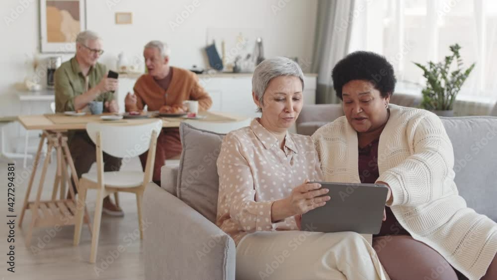 Medium long of senior Asian and African female friends sitting on couch in living room, watching something on tablet computer, smiling, talking in foreground of men communicating Medium long of senior Asian and African female friends sitting on couch in living room, watching something on tablet computer, smiling, talking in foreground of men communicating