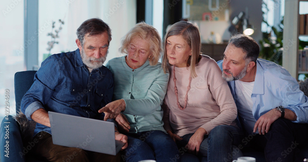 Two caucasian married couples staying in living room using computer pc laptop browsing news shopping e-commerce buying tickets communicating.