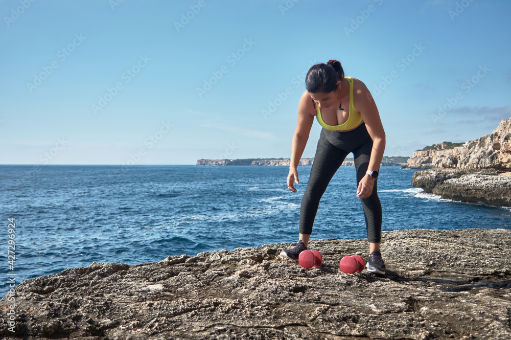 FITNESS LATIN WOMAN IN SPORTS SET TRAINING WITH ELASTIC BAND, WEIGHTS, GYM EXERCISES, IN FRONT OF THE WATER.