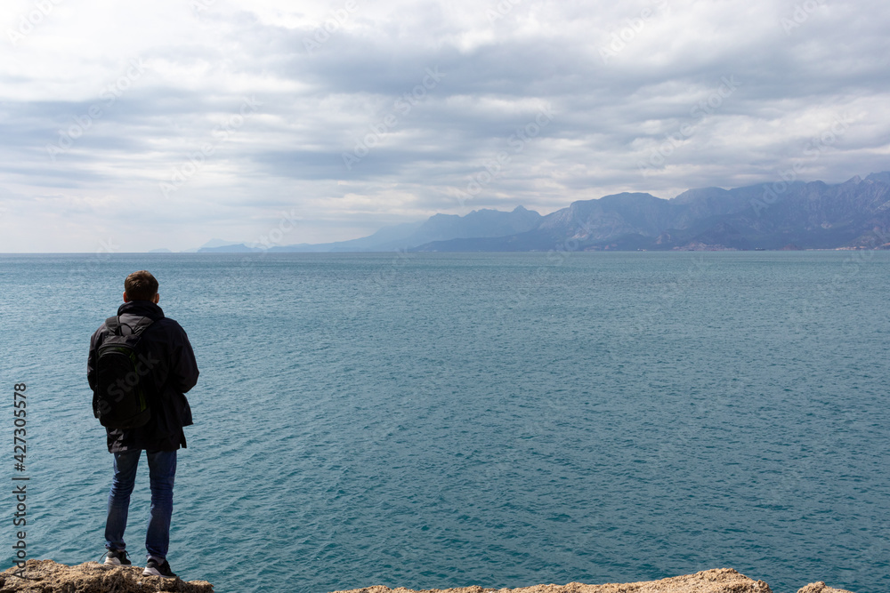 Young casual man traveller with backpack stands on high stone shore and looking in the sea distance. Web banner for travel and exploring the natural world.