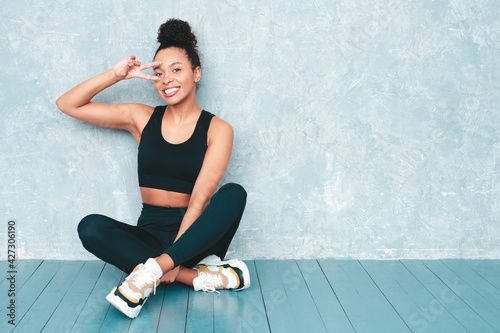 Fitness smiling black woman in sports clothing with afro curls hairstyle.She wearing sportswear. Young beautiful model with perfect tanned body.Female sitting in studio near gray wall after training