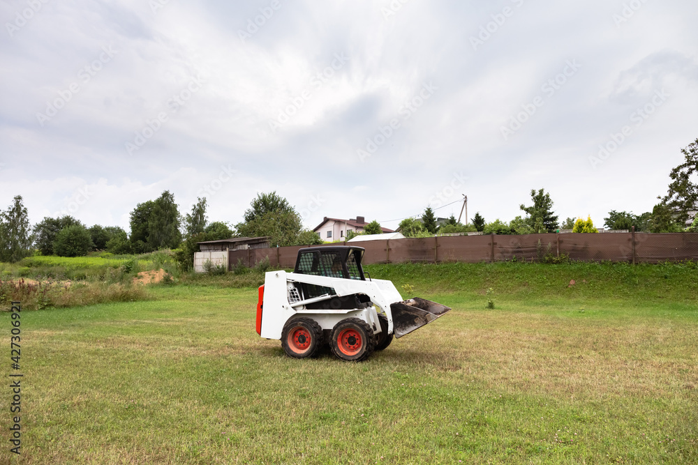 A skid steer loader clears the site for construction. Land work by the territory improvement. Machine for work in confined areas. Small tractor with a bucket for moving soil and bulk materials.