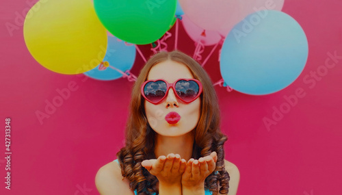 Portrait of beautiful woman blowing her red lips sending air kiss with colorful balloons on a pink background