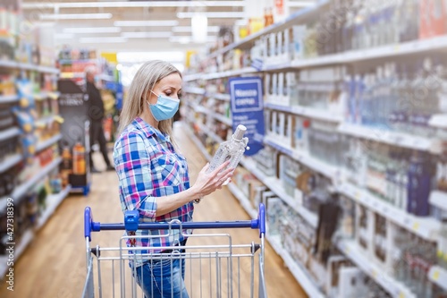 Woman in medical mask reading the label of vodka bottle in liquor store or alcohol section of supermarket.
