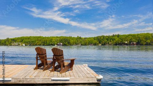 Two Adirondack chairs on a wooden dock facing a lake in Muskoka, Ontario Canada during a sunny summer morning. Cottages are nested between trees across the water.