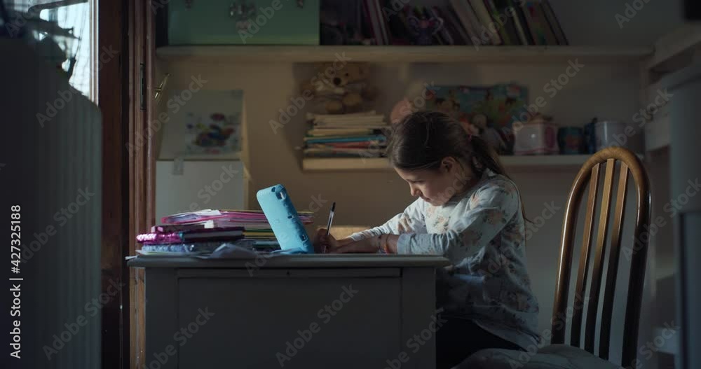 Authentic shot of little girl pupil is writing in notebook at desk while having online lecture with tablet with a teacher in virtual classroom during distance education in her room at home. Covid Authentic shot of little girl pupil is writing in notebook at desk while having online lecture with tablet with a teacher in virtual classroom during distance education in her room at home. Covid