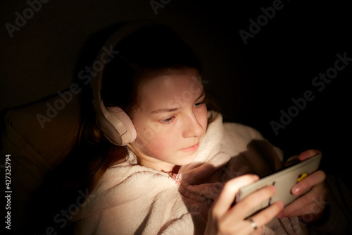 a teenage girl in wireless headphones at night in bed with a smartphone in her hands listens to streaming music