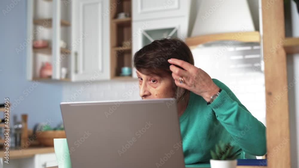 Happy elderly woman excitedly communicating by video call using laptop at home in kitchen. Mature middle-aged grandmother talks online with her family and grandchildren. Distance communication concept Happy elderly woman excitedly communicating by video call using laptop at home in kitchen. Mature middle-aged grandmother talks online with her family and grandchildren. Distance communication concept