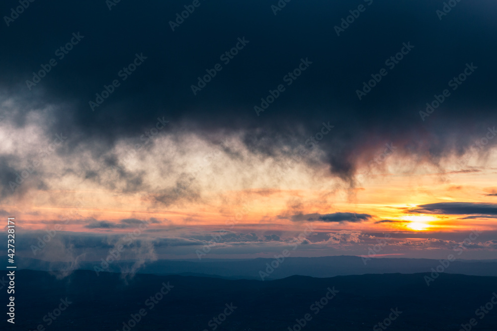 Sunset with sun hidden behind clouds over mountains and valley, with very low and close clouds