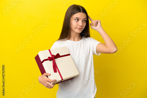 Little girl holding a gift over isolated yellow background having doubts and thinking