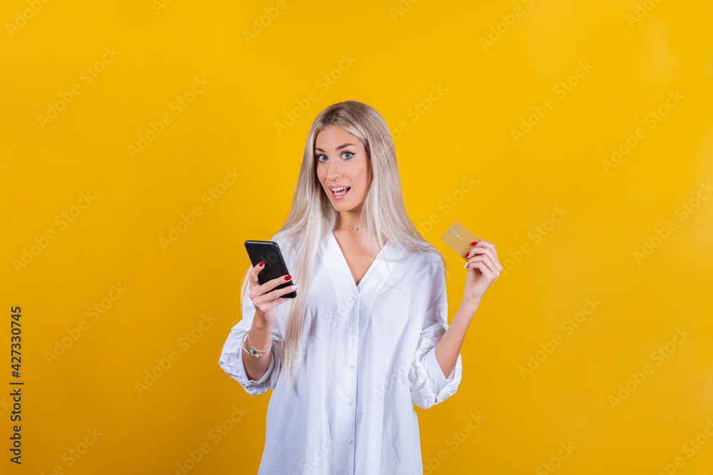 Portrait of a happy young blonde girl showing debit card while holding cell phone isolated on yellow background - Close up of shocked young woman using smartphone holding credit card Portrait of a happy young blonde girl showing debit card while holding cell phone isolated on yellow background - Close up of shocked young woman using smartphone holding credit card
