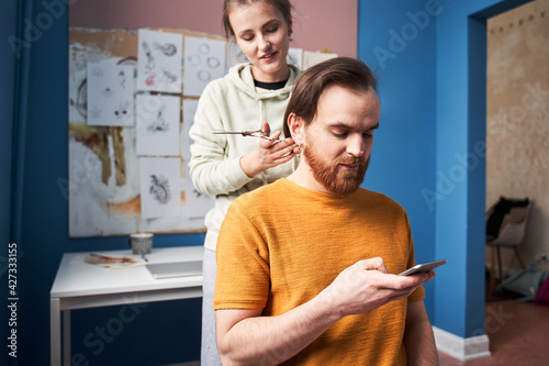 Woman holding scissors and looking at her man while cutting his hair at the kitchen