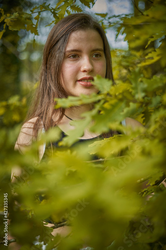 Light-skinned young woman with long hair, black top and necklace among the leaves and branches of an oak tree. girl in nature. teenage girl among the branches of a green oak tree smiling and pensive.