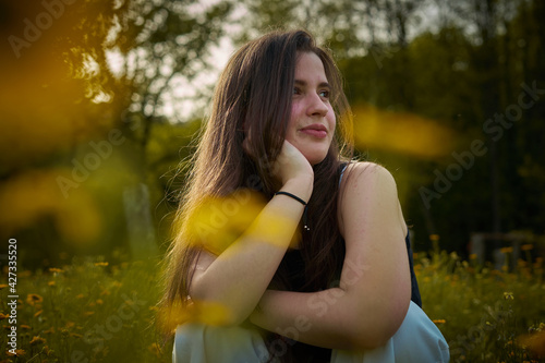Light-skinned young woman with long hair, black top and white pants sitting in field of yellow flowers smiling and thinking. girl in nature. teenager with daisies thinking and smiling. eco person