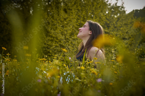 Light-skinned young woman with long hair, black top and white pants sitting in field of yellow flowers smiling and thinking. girl in nature. teenager with daisies thinking and smiling. eco person