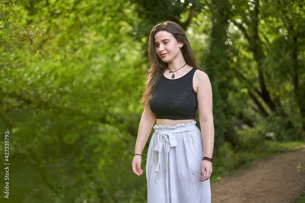 Young girl with dark hair and light skin in a black top, slouchy white pants, black and white sneakers and necklace `walking along a dirt path in lush green forest. woman in nature. teenager hiking Young girl with dark hair and light skin in a black top, slouchy white pants, black and white sneakers and necklace `walking along a dirt path in lush green forest. woman in nature. teenager hiking