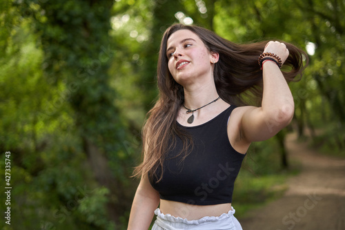 girl with long dark hair, light skin, black top and white pants placing her hair with her hand in the forest. woman combing her hair in nature teenage girl pushing hair out of face