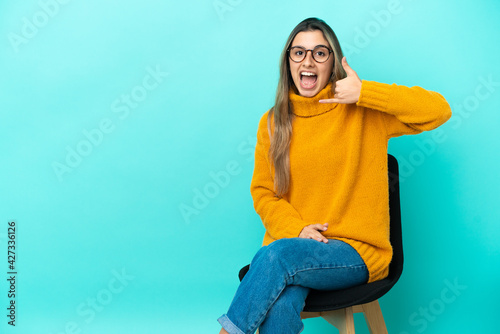 Young caucasian woman sitting on a chair isolated on blue background making phone gesture. Call me back sign © luismolinero