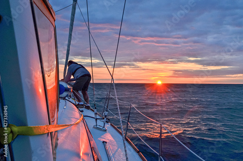 A man secures his sailboat as the setting sun bathes the sea, boat, and sky in warm red orange light.  Copy space 