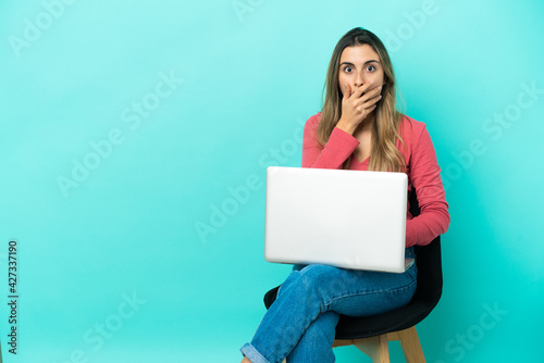 Young caucasian woman sitting on a chair with her pc isolated on blue background surprised and shocked while looking right © luismolinero