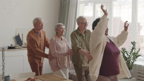 Slow motion medium long of happy multiethnic senior friends dancing conga at home around dining table photo