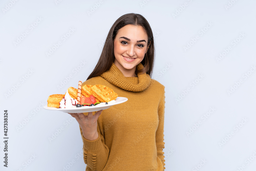 Young caucasian woman holding waffles isolated on blue background with happy expression Young caucasian woman holding waffles isolated on blue background with happy expression