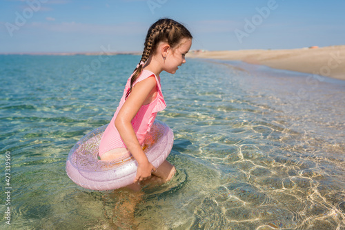 Happy cute tollder girl 5-6 years old in bright pink swimsuit with swim tubes ring going into the sea for swimming on sunny day. Kids having fun at the beach. Family vacation with children concept photo