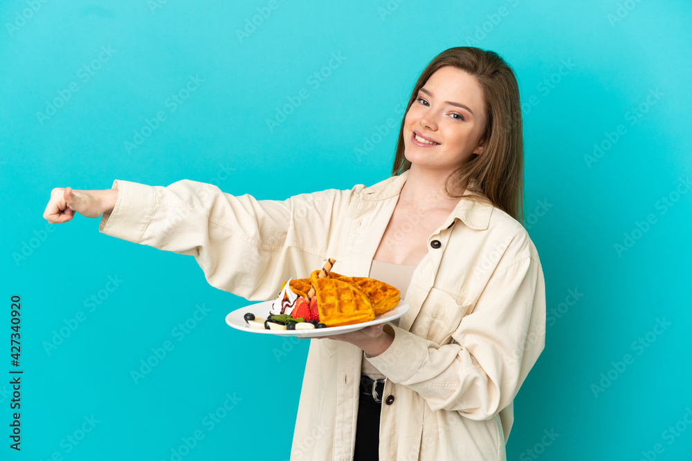 Teenager girl holding waffles over isolated blue background giving a thumbs up gesture
