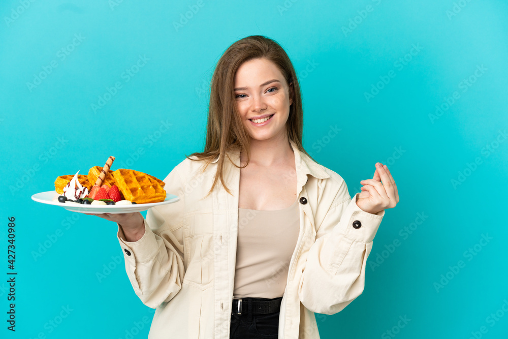 Teenager girl holding waffles over isolated blue background making money gesture Teenager girl holding waffles over isolated blue background making money gesture