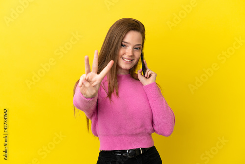 Teenager girl using mobile phone over isolated yellow background happy and counting three with fingers