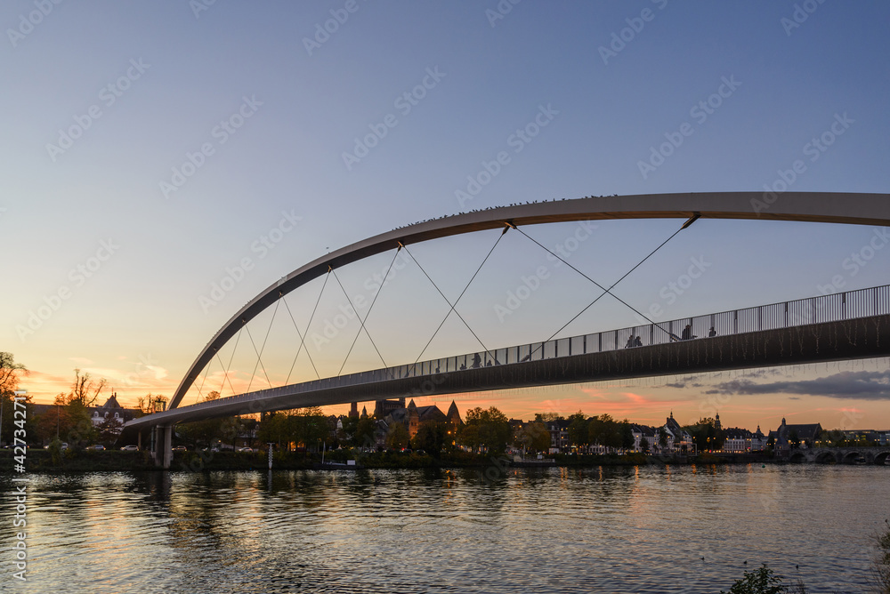 Outdoor scenery on riverside of Meuse river and Hoge Brug, Modern pedestrian bridge, during sunset time and dramatic twilight sky in Maastricht, Netherlands.  