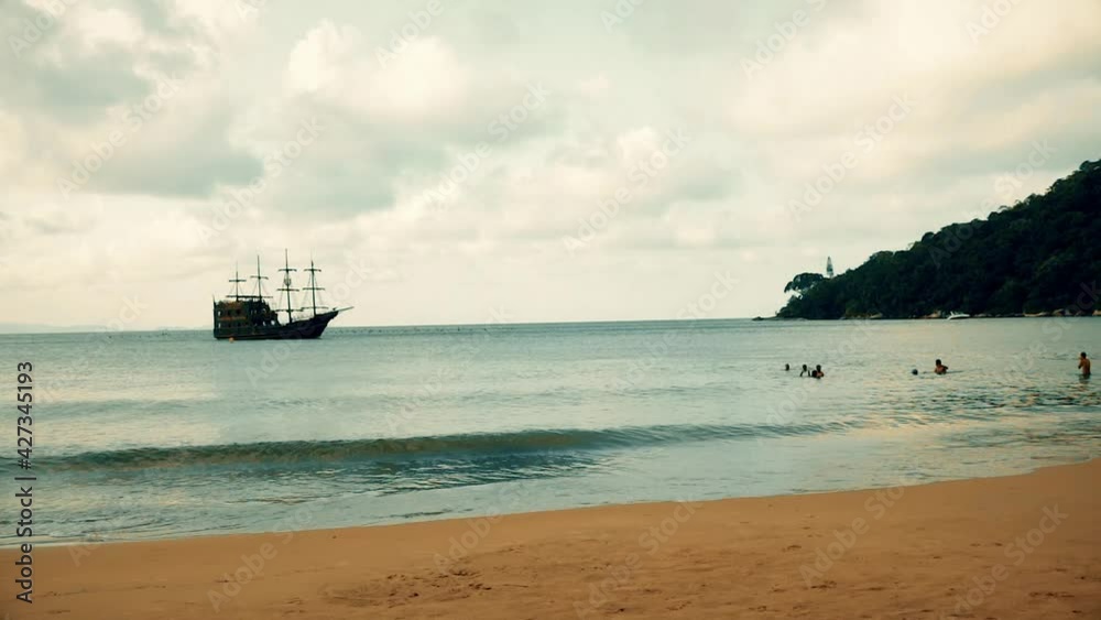 A medieval ship is getting closer to the beach to dock on the port on a cloudy day with people on the water. Old ship similar to Columbus vessel, exploring america with navigator skills on calm sea A medieval ship is getting closer to the beach to dock on the port on a cloudy day with people on the water. Old ship similar to Columbus vessel, exploring america with navigator skills on calm sea