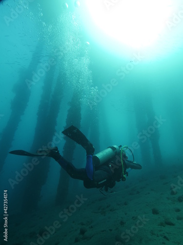 A scuba diver diving underwater near an underside pier with sunlight in the water.