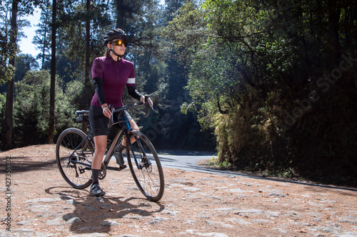 Joven ciclista descansando con su bicicleta al lado de la carretera , usando casco y lentes del sol, en medio del bosque. deporte al aire libre