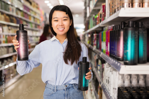 Portrait of attractive Asian girl holding and choosing plastic bottle of shampoo in cosmetics shop