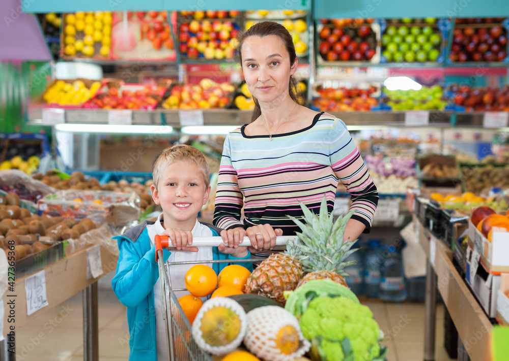 Happy cheerful little boy with his positive pleasant mother choosing fresh fruits and vegetables at supermarket