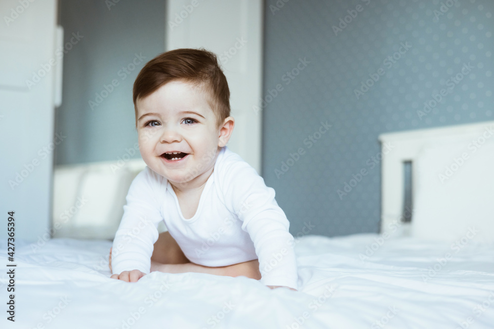 A portrait of a cute toddler baby boy in a white bodyuit seating on bed and laughing.Happy baby face look at the camera, in bedroom at home A portrait of a cute toddler baby boy in a white bodyuit seating on bed and laughing.Happy baby face look at the camera, in bedroom at home