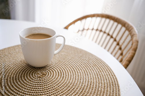 White cup of coffee on wicker place mat. Background with white table and rattan chair. Boho style kitchen. Copy space. Hygge. 