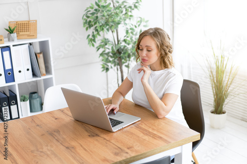 Business woman using laptop sitting near desk white office interior with houseplant looking at camera