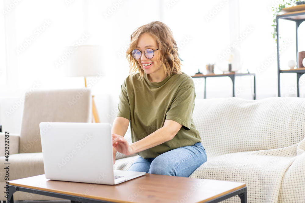 Woman glasses using laptop computer while sitting sofa with big window on background at home interior Woman glasses using laptop computer while sitting sofa with big window on background at home interior