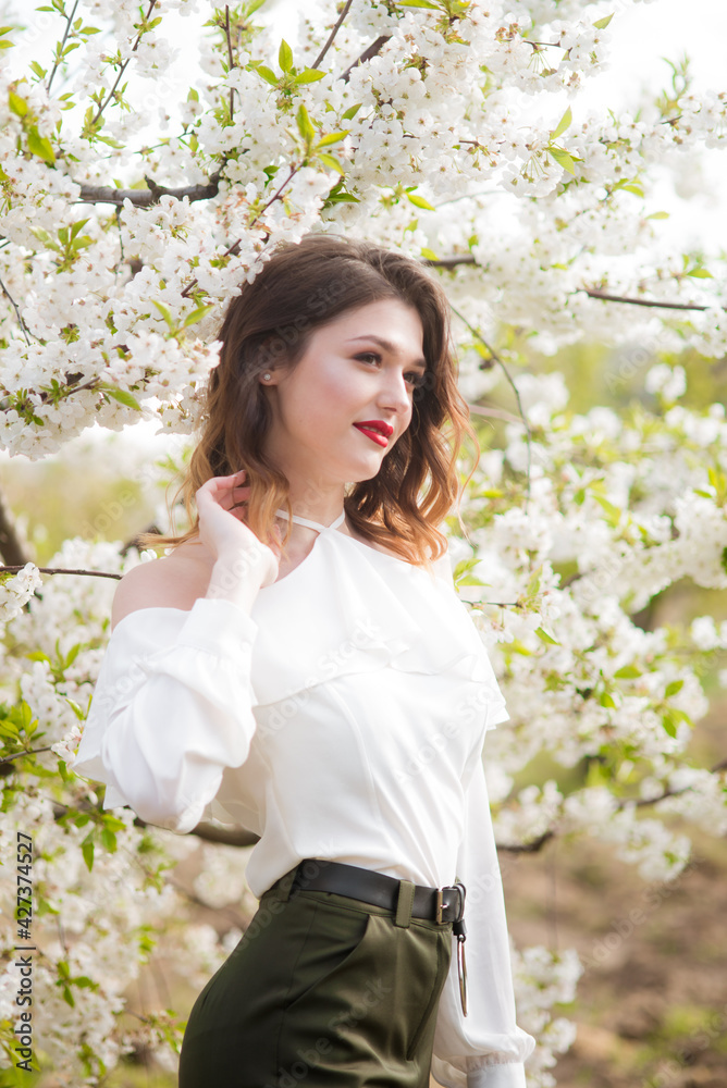 Lovely young girl in a white romantic silk blouse in spring in a blooming cherry garden in sunny weather. Spring and Fashion. Spring blooming gardens Lovely young girl in a white romantic silk blouse in spring in a blooming cherry garden in sunny weather. Spring and Fashion. Spring blooming gardens
